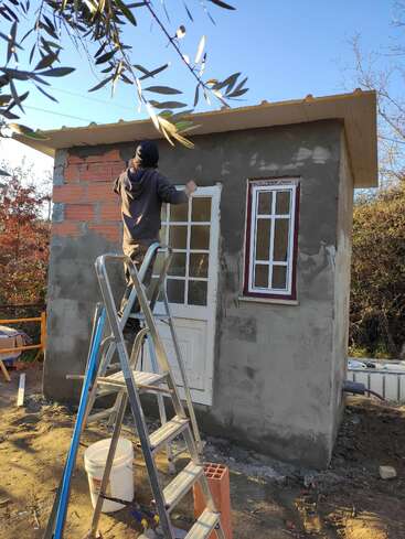 Une personne se tient sur une échelle devant un petit bâtiment inachevé, travaillant sur le mur extérieur. Des outils et des matériaux de construction sont éparpillés sur le site.