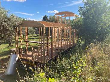 Une structure en bois partiellement construite est posée sur une remorque, entourée de verdure et de soleil, sous un ciel bleu parsemé de nuages. Des matériaux de construction se trouvent à proximité.