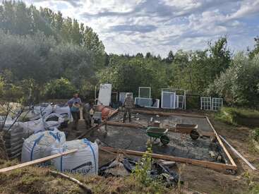 Trois personnes travaillent sur les fondations d'une maison dans une clairière, à l'aide d'une bétonnière. De grands sacs, des brouettes et des cadres de fenêtres sont éparpillés sur le site.