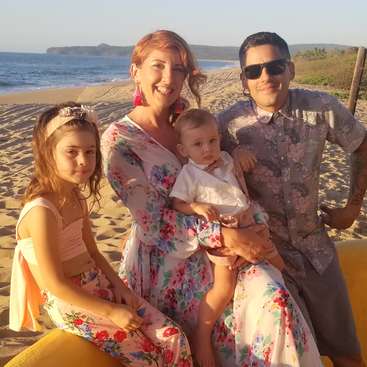 La imagen muestra a una familia de cuatro miembros posando en una playa de arena, con la madre sosteniendo a un bebé y el padre de pie a su lado, vestidos con trajes florales a juego.