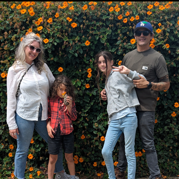The image depicts a family of four posing in front of a vibrant orange flower wall, with the adults standing on either side of two children.