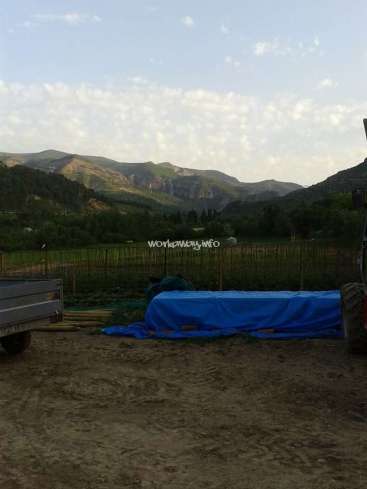 The image depicts a serene landscape with a mountain range in the background, featuring a blue tarp, a trailer, and a tractor in the foreground.