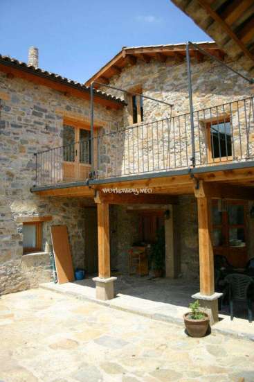 The image depicts a rustic stone house with a balcony, featuring a wooden door and windows, set against a clear blue sky.