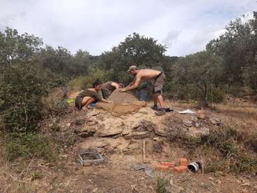 The image depicts three individuals constructing a stone structure in a rural setting, surrounded by trees and bushes, with a cloudy sky above.