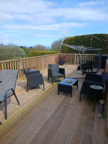 A spacious wooden deck with black outdoor furniture, a hanging chair, potted plants, and a rotary clothesline. Surrounded by lush greenery and blue sky.