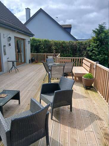 A spacious wooden deck with black outdoor seating, a round glass table, potted plants, surrounded by a wooden fence, tall hedges, and neighboring houses under cloudy skies.