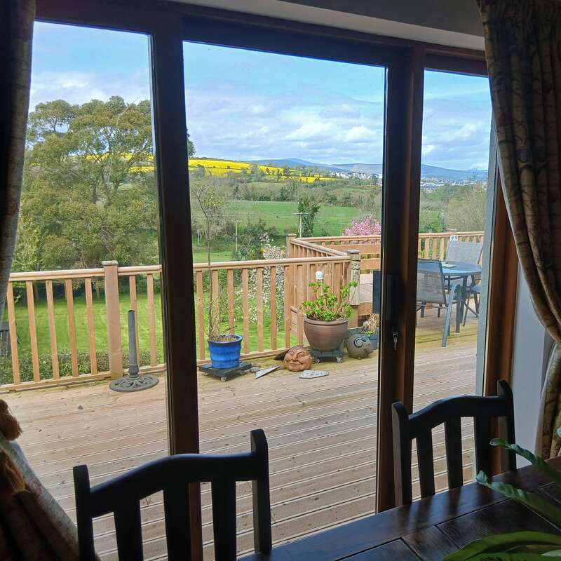 A peaceful countryside view through large glass doors reveals a wooden deck with potted plants, outdoor furniture, green fields, trees, and distant yellow fields.