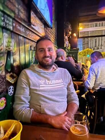 Ein lächelnder Mann in einem grauen Hummel-Hoodie sitzt mit einem Drink an einem gemütlichen Bartisch. Bunte, verzierte Wände und andere Gäste prägen die lebhafte Atmosphäre.