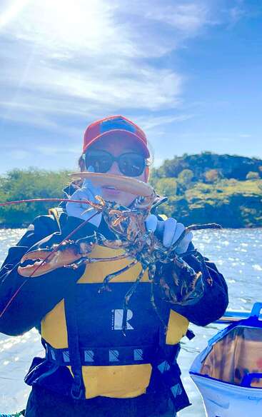 La imagen muestra a una persona con chaleco salvavidas y una langosta en la mano, de pie en un barco con una pintoresca masa de agua y árboles de fondo bajo un cielo azul.