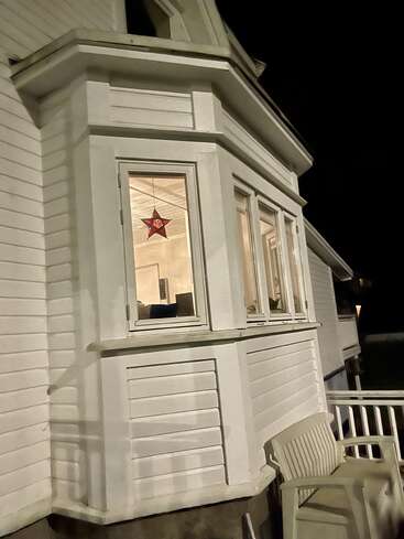 La imagen representa una casa blanca con una ventana mirador de tres hojas, adornada con una estrella roja que cuelga de la hoja central, sobre un cielo nocturno oscuro.