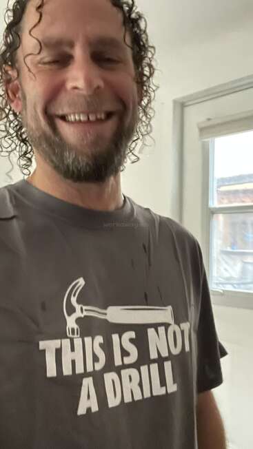 Un homme souriant aux cheveux bouclés et à la barbe porte une chemise humoristique avec un marteau et les mots "THIS IS NOT A DRILL" à l'intérieur d'une pièce.
