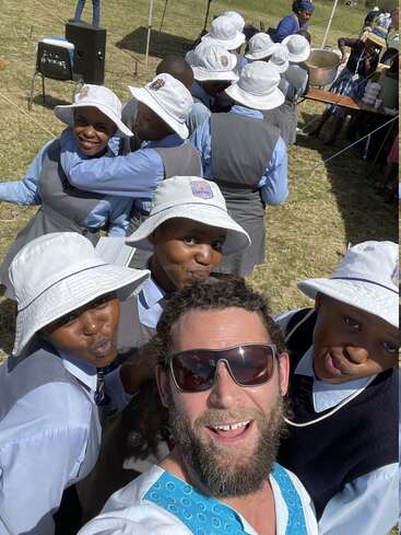 Un homme barbu prend un selfie joyeux avec un groupe d'écoliers en uniformes et chapeaux blancs. Ils sont en plein air, souriants et profitent d'une journée ensoleillée.