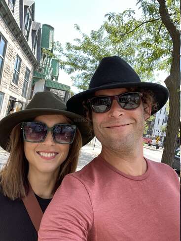 Un couple souriant prend un selfie en plein air. Tous deux portent des lunettes de soleil foncées et des chapeaux élégants. Derrière eux, de vieux bâtiments et des arbres feuillus complètent la scène lumineuse et joyeuse.