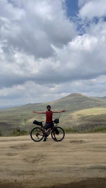 Un cycliste se tient debout, les bras tendus, à côté d'un vélo emballé sur un chemin de terre, entouré de vastes collines vertes et sous un ciel dramatique et nuageux.