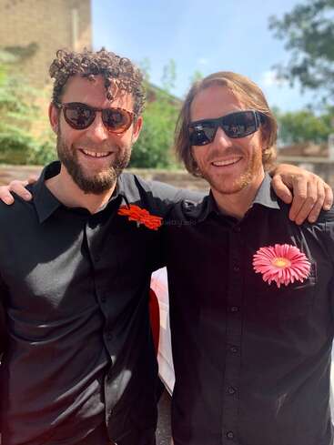Deux hommes souriants portant des chemises noires et des lunettes de soleil se tiennent ensemble en plein air, chacun avec une fleur sur la poitrine. Ils ont l'air heureux et à l'aise, profitant de la lumière du soleil.