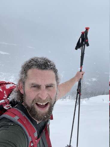 Un homme barbu, couvert de neige, sourit avec enthousiasme en tenant des bâtons de ski dans un paysage montagneux enneigé, portant un sac à dos rouge, profitant d'une aventure hivernale froide.