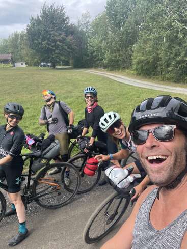 Cinq cyclistes, tous casqués et portant des lunettes de soleil, sourient joyeusement pour un selfie. Ils sont en plein air, le long d'une route, entourés d'arbres verts et de champs herbeux.
