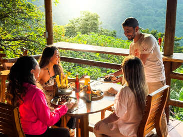 Trois femmes sont assises à une table en bois, savourant un repas et un verre en plein air, face à une magnifique vue sur la forêt verdoyante. Un homme souriant leur sert à manger sous les rayons du soleil.