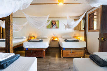 Cette photo montre une chambre commune chaleureuse équipée de cinq lits simples, de moustiquaires, de meubles en bois, d'un éclairage tamisé, de serviettes soigneusement pliées et d'une photo joyeuse d'un enfant.