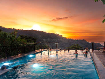 Les gens se détendent dans une piscine à débordement surplombant des collines verdoyantes, sous un coucher de soleil flamboyant, tandis que le ciel aux teintes orange et jaune vif crée une atmosphère sereine et pittoresque.