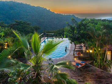 Ce havre de paix tropical offre une piscine accueillante entourée d'une végétation luxuriante, une terrasse en bois, des hamacs, des guirlandes lumineuses et, en toile de fond, les montagnes se découpant sur le ciel au moment d'un coucher de soleil serein.