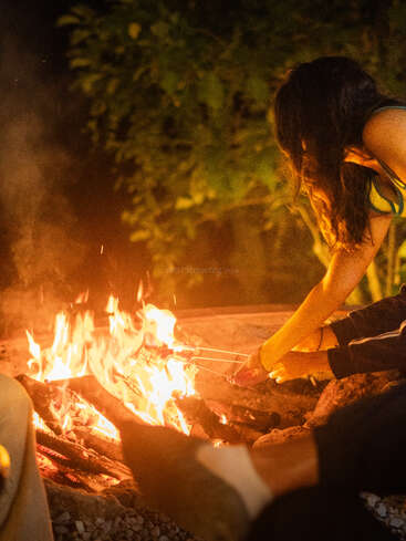 Deux personnes sont assises près d'un feu de camp qui crépite dans la nuit, en train de faire griller des marshmallows au-dessus des flammes, entourées de rochers et d'arbres, profitant d'une ambiance chaleureuse en plein air.
