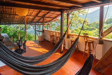 Une terrasse couverte avec un sol en terre cuite, des hamacs confortables, des tabourets de bar en bois, de grandes tables en bois et une vue imprenable sur les montagnes verdoyantes, baignée par la douce lumière naturelle du soleil.