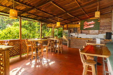 Cette image montre un bar en plein air à l'ambiance chaleureuse et rustique, avec des tables et des chaises en bois, un éclairage tamisé, une enseigne au néon, une végétation luxuriante à l'extérieur et une atmosphère décontractée et accueillante.
