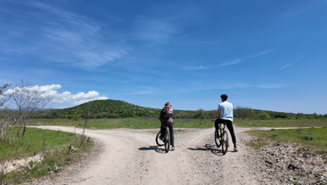 Zwei Radfahrer halten an einer Kreuzung auf einem Feldweg unter strahlend blauem Himmel an, umgeben von grünen Feldern und einem Hügel in der Ferne, und genießen die Natur.