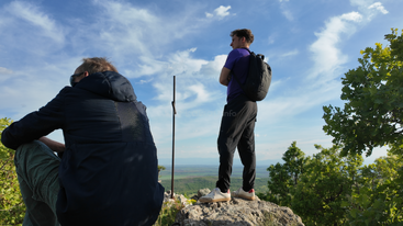 Zwei Männer stehen auf einem felsigen Hügel, umgeben von Grün, blicken auf eine weite Landschaft unter einem blauen Himmel mit vereinzelten Wolken und genießen die malerische Aussicht.