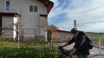 Ein lächelnder Mann mit Helm fährt mit dem Fahrrad vor einem weißen Haus mit Garten vorbei. Der Himmel ist teilweise bewölkt, was auf einen angenehmen Tag hindeutet.