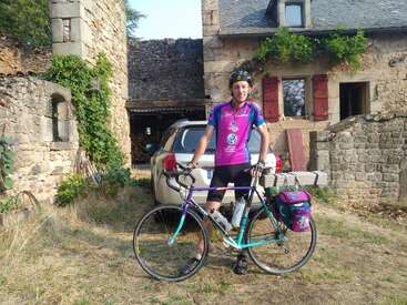 Ein Radfahrer in einem lila Trikot und mit Helm steht an einem sonnigen Tag mit einem Tourenrad vor einem rustikalen Steinhaus und einem geparkten Auto.