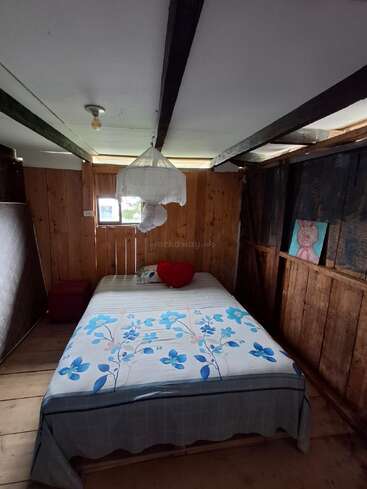 Una acogedora habitación rústica de madera cuenta con una cama con ropa de cama azul con motivos florales, un cojín rojo en forma de corazón, mosquitera, iluminación mínima, una pequeña ventana, colchón y un cuadro.