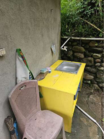 An outdoor washing area features a yellow sink unit, faucet, scrubbing brush, broom, plastic chair, towel, stone wall, and surrounding green plants. Rustic and practical.