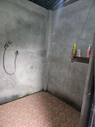 This image shows a simple, bare shower with concrete walls, patterned floor tiles, a showerhead, and a small shelf holding toiletries under a corrugated roof.