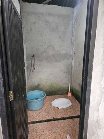 This is a simple, traditional squat toilet with a plastic water tub, a dipper, a toilet brush, tiled floor, and unfinished concrete walls, inside a small room.