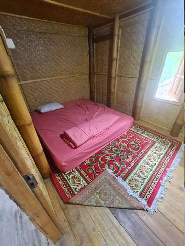 This image shows a cozy bamboo room with woven walls, a simple mattress with pink bedding, a single pillow, and a colorful, patterned rug on wooden flooring.