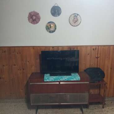 A imagem retrata um suporte de TV moderno de meados do século com uma televisão de tela plana, uma pequena mesa e objetos decorativos pendurados na parede contra uma parede com painéis de madeira e fundo branco.