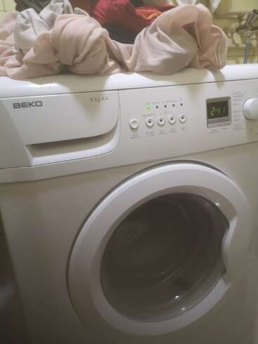 Uma máquina de lavar branca da Beko exibe um cronômetro digital com a indicação 2:41. Várias peças de roupa estão empilhadas em cima, prontas para serem lavadas. Configuração da lavanderia.