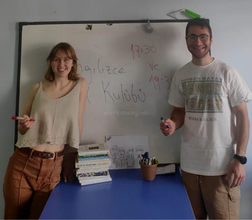 Dois jovens adultos, sorridentes, estão ao lado de um quadro branco em uma sala de aula. Livros e marcadores sobre a mesa. A placa acima diz "GVZ Dil Kulübü" em turco.