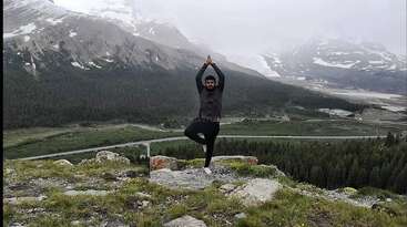 A person is performing yoga outdoors, standing in a seated position with legs extended, arms raised, and hands together, wearing a white shirt and pants, in a natural setting with greenery and trees.