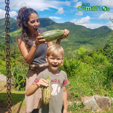 Una mujer y un niño posan alegremente al aire libre, sosteniendo calabacines frescos. Las exuberantes colinas verdes y el cielo azul crean un fondo hermoso y vibrante. Logotipo: «Eco Caminhos» arriba.
