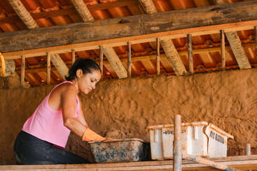 Una mujer con guantes y una camiseta rosa sin mangas está construyendo una pared de barro bajo un techo de tejas, alisando cuidadosamente la superficie con las manos. Herramientas de construcción cerca.
