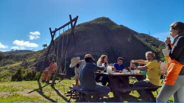 Un grupo de personas está sentado en una mesa de picnic al aire libre, disfrutando de comida y bebida con las montañas al fondo. Se ve un columpio y un cielo azul claro.
