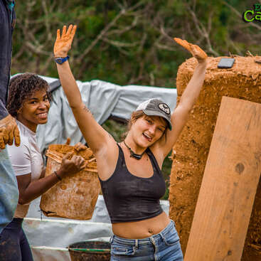 Dos mujeres trabajan alegremente al aire libre con arcilla o barro, sonriendo y levantando los brazos mientras participan en un proyecto práctico de construcción o edificación rodeadas de naturaleza.