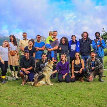 A group of people, including a child and a dog, pose outdoors on green grass with mountains and a blue sky in the background, smiling happily.
