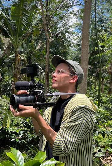Un homme portant des lunettes et une casquette filme dans une forêt luxuriante avec une caméra professionnelle, entièrement concentré sur la nature qui l'entoure, au milieu d'une végétation éclatante.