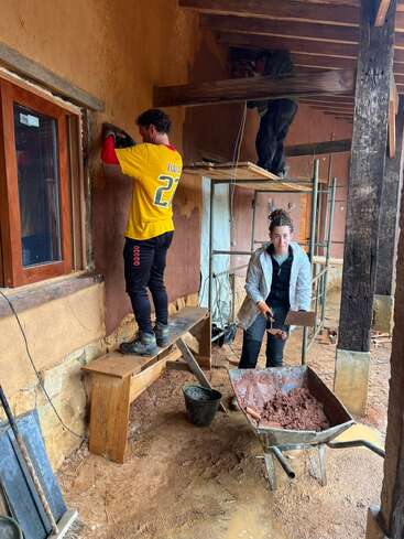 Trois personnes travaillent sur un bâtiment rustique : l'une enduit un mur, une autre mélange des matériaux dans une brouette, tandis que la troisième travaille en hauteur sur un échafaudage.