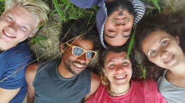 Cinq amis sont allongés en cercle sur l'herbe et sourient à la caméra. Ils ont l'air heureux et détendus, profitant ensemble d'un moment agréable en plein air.