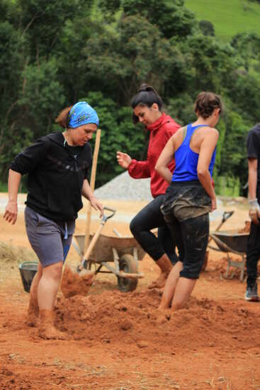 Tres mujeres pisan con los pies descalzos sobre tierra rojiza, removiéndola, quizá para la construcción. Cerca hay carretillas y palas. El fondo está cubierto de frondosos árboles y hierba.